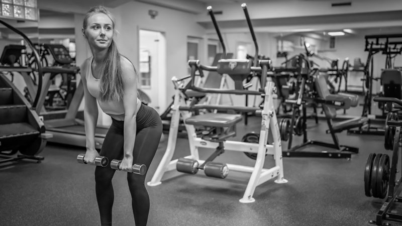 Mujer realizando peso muerto rumano con mancuernas en un gimnasio, rodeada de máquinas de entrenamiento de fuerza.