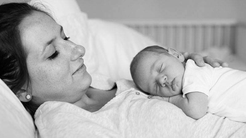 Madre descansando en la cama con su bebé recién nacido dormido sobre el pecho durante el periodo de posparto.
