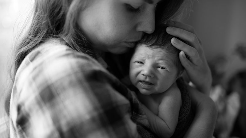 Madre sosteniendo y protegiendo a su bebé recién nacido en brazos durante el posparto, en un entorno interior tranquilo.
